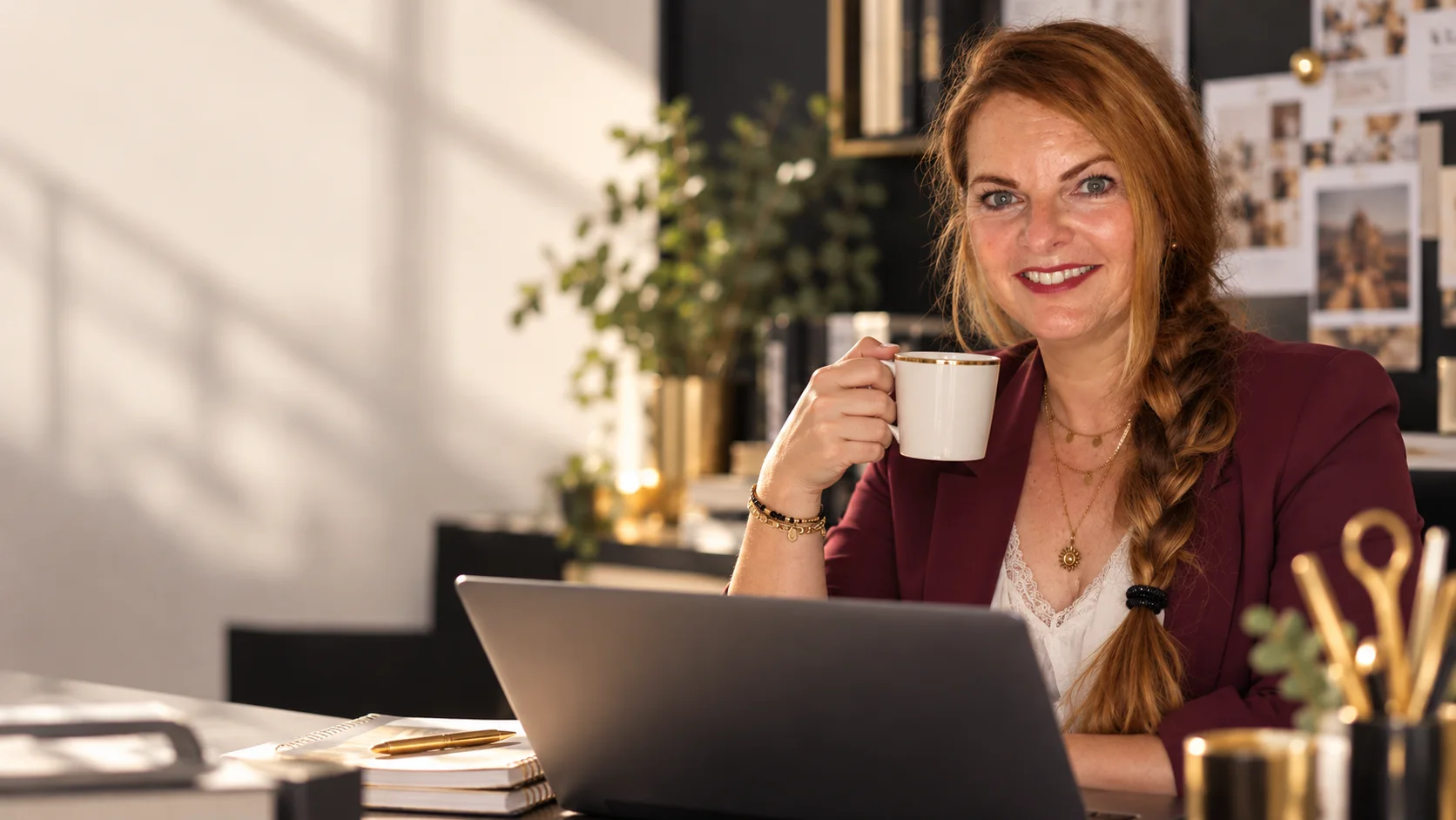 Denise Dobra im burgunderfarbenen Blazer mit Kaffeetasse vor ihrem Laptop